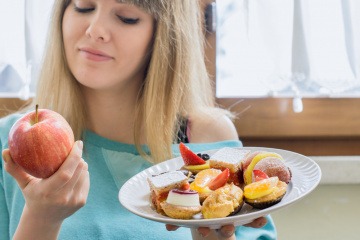 Eine Frau hält einen Apfel und ein Teller mit Süßem und entschließt sich für eine Ernährungsumstellung