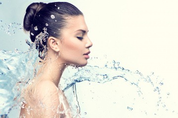 Eine Frau mit jochgesteckten Haaren im Profil. Ein Schwall Wasser bewegt sich gerade in Richtung ihr Gesicht