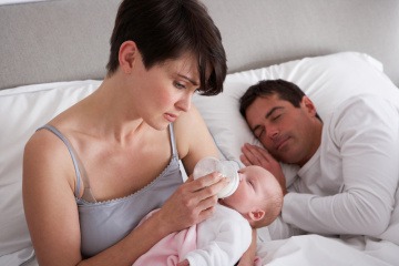 Eine Frau füttert ihr Baby mit der Flasche, der Mann schläft daneben