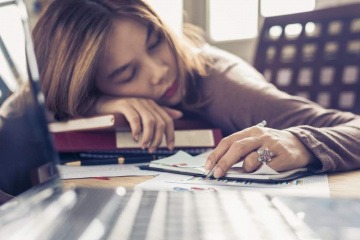 Eine Frau liegt mental erschöpft am Schreibtisch über Büchern und Unterlagen