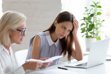 Eine Frau sitzt genervt am Tisch, während ihre Kollegin einen langweiligen Monolog hält.