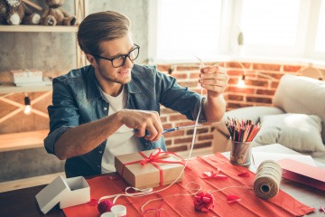 Geschenke originell verpacken Tipps - junger Mann verpackt Präsente auf Tisch.