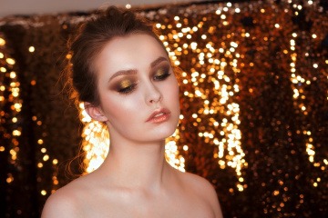 Eine Frau steht vor einem goldenen Hintergrund und trägt goldenes Glitzer-Makeup