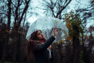 Eine Frau wirkt grundlos traurig und berührt die Innenseite eines transparenten Regenschirms an der Stelle, an der außen ein Laubblatt draufgehalten ist.