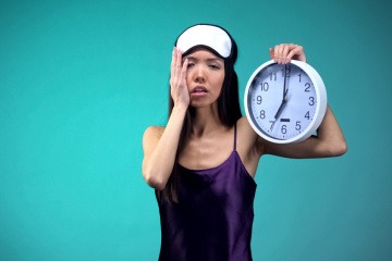 Innere Uhr umstellen - müde wirkende Frau im Nachthemd hält eine Wanduhr hoch.