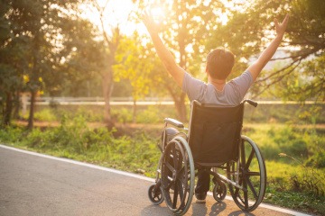 Selbstbestimmtes Leben führen - ein Mann im Rollstuhl, bei Sonnenaufgang in einem Park, hebt die Arme in Siegerpose.