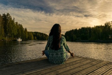 Eine Frau sitzt auf einem Holzsteg am Wasser und meditiert