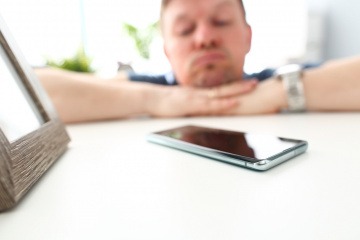 Ein Handy liegt auf dem Tisch. Ein nachdenklicher Mann im Hintergrund wartet auf einen Anruf oder eine Nachricht einer Frau.