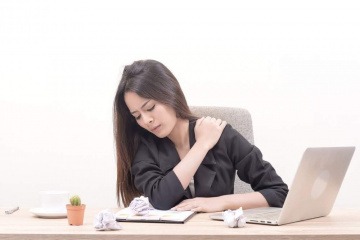 Eine am Schreibtisch sitzende Frau greift sich mit schmerzverzerrtem Gesicht an die Schulter. Rückenschmerzen, Homeoffice und eine fehlende Routine bei der Heimarbeit hängen oft miteinander zusammen.