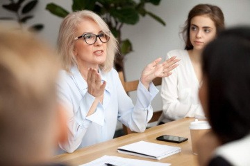 Eine ältere Frau diskutiert mit anderen Menschen an einem großen Tisch.