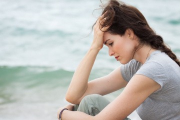 Eine Frau sitzt auf einer Düne am Strand, der Kopf ist nach unten gesenkt, ihr Ausdruck wirkt traurig und ratlos.