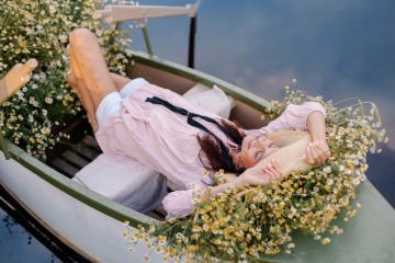 Frau mit Hut liegt im Sommer in einem mit Blumen ausgelegten Boot und lässt sich treiben.