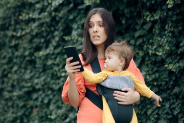 Stress durch Handy - Mutter mit Baby im Arm blickt gestresst auf ihr Smartphone.