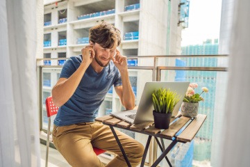 Ein junger Mann sitzt am Balkon und hält sich die Ohren zu