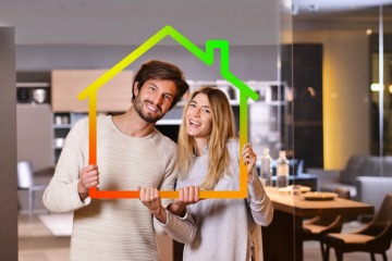 Strom sparen zuhause - junges Paar hält ein kleines Haus-Symbol hoch, dessen Umrisse in grün, gelb und rot gefärbt sind.