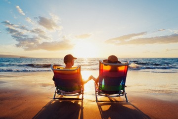 Paar beobachtet Sonnenuntergang am Strand und hält dabei Händchen.