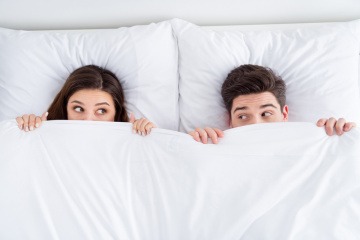 Paar liegt verklemmt im Bett, die Decke bis zum Gesicht gezogen