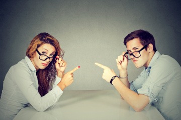 Ein Paar sitzt am Tisch, beide zeigen mit dem Finger auf ihr Gegenüber. Es geht um Vorwürfe, Beziehung und ähnliches.