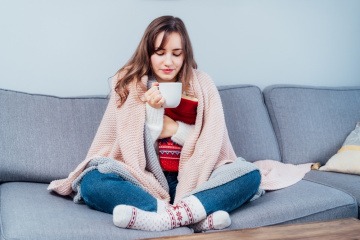 Wärmekissen selber machen - Frau sitzt mit Wärmekissen und Decke über den Schultern auf ihrem Sofa und trinkt Tee.