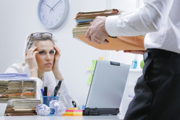 Zu viel arbeiten - Frau am Schreibtisch ist gestresst, ihr Schreibtisch voller Akten, Chef bringt neue. 